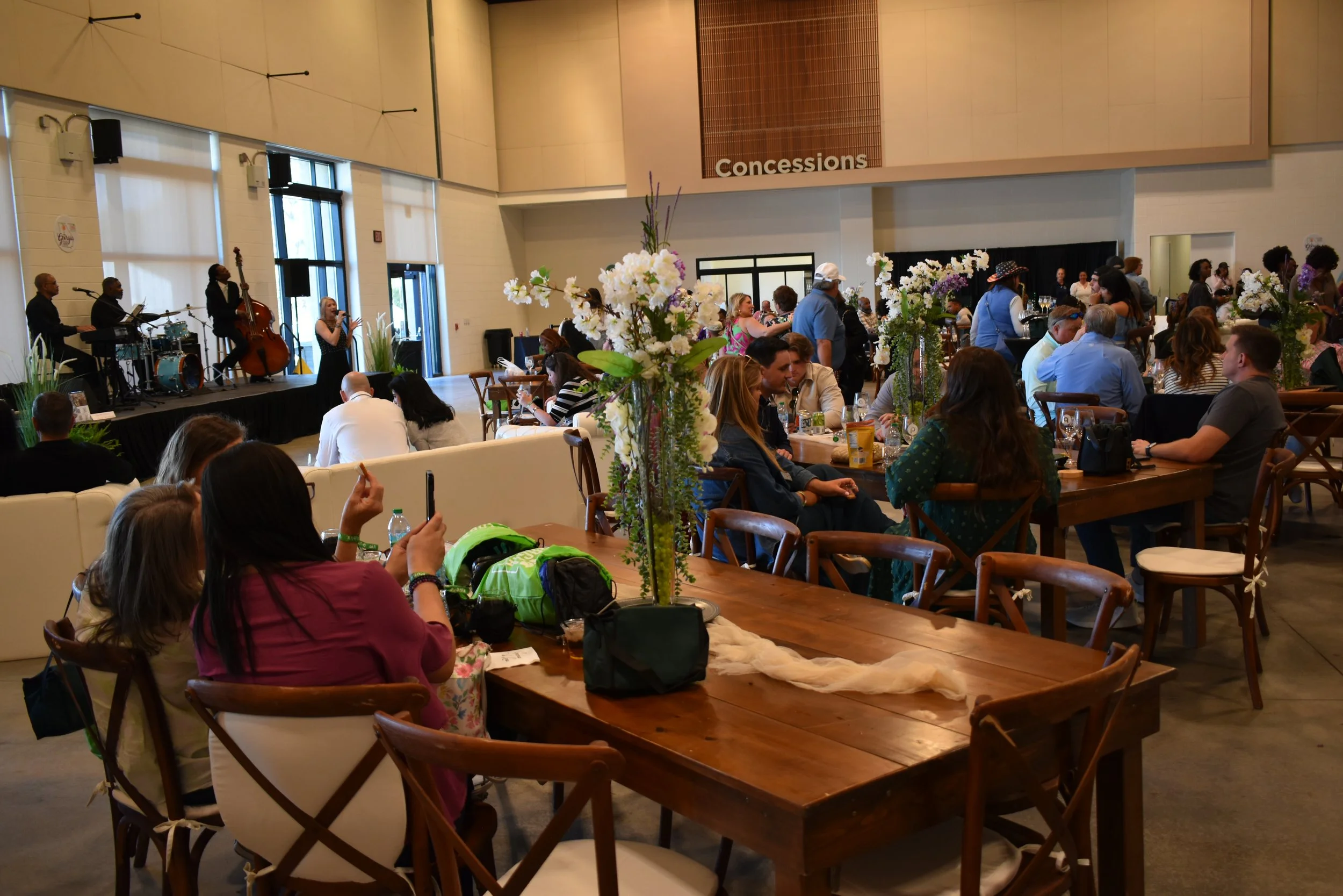 Indoor event with live band playing on stage, guests seated at tables with floral centerpieces, some taking photos, large windows in background.