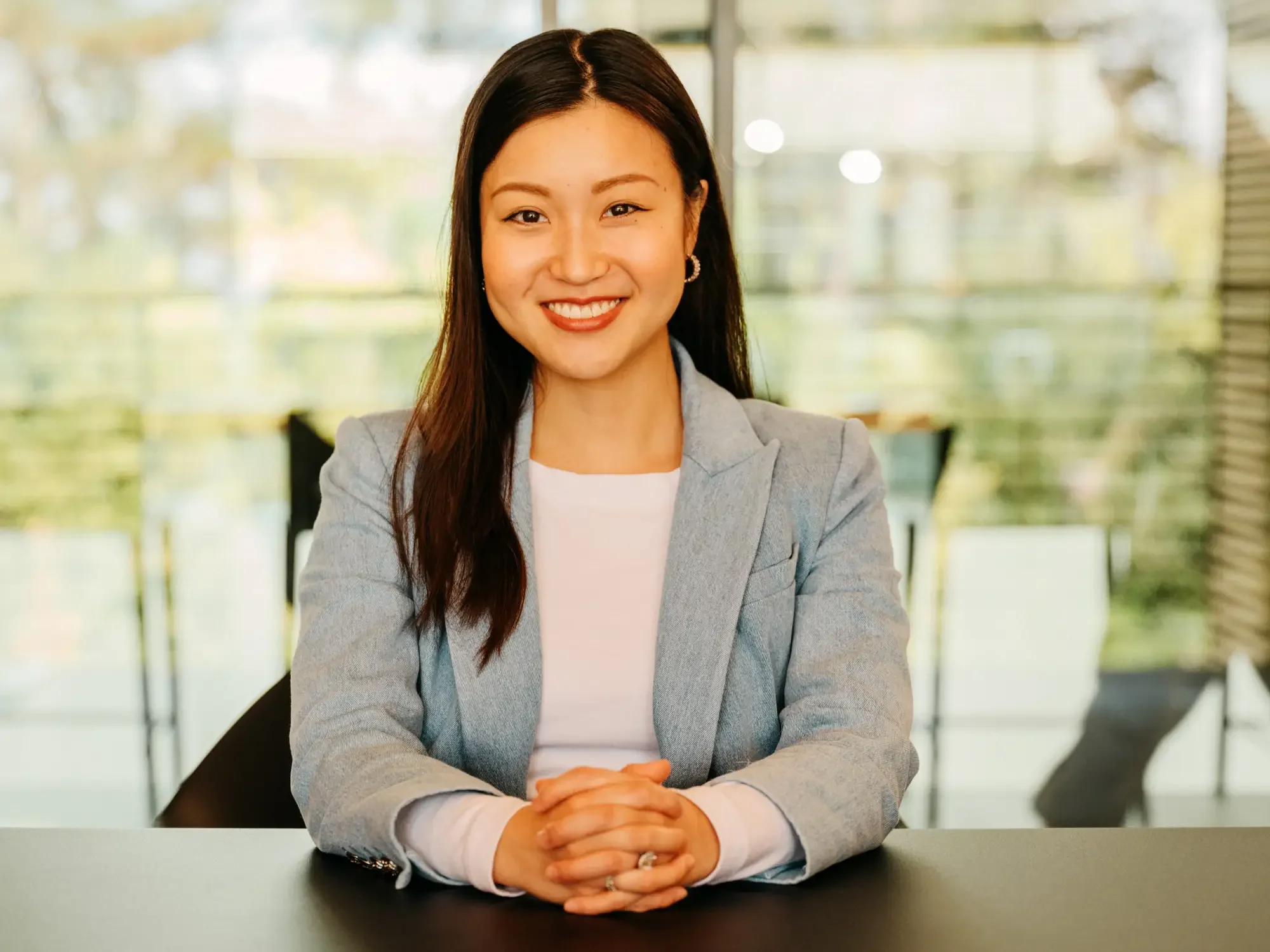 A woman with long dark hair, wearing a light gray blazer and a white top, sitting at a table with her hands clasped, smiling at the camera in a modern office setting with large windows and blurred greenery in the background.