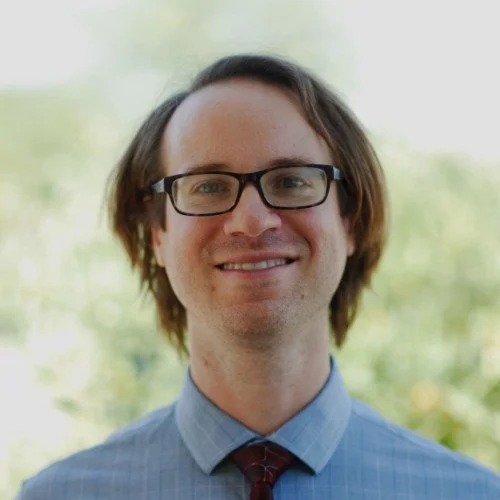 A young man with shoulder-length brown hair, wearing black glasses, a light blue collared shirt, and a red tie, smiling outdoors with blurred green foliage in the background.