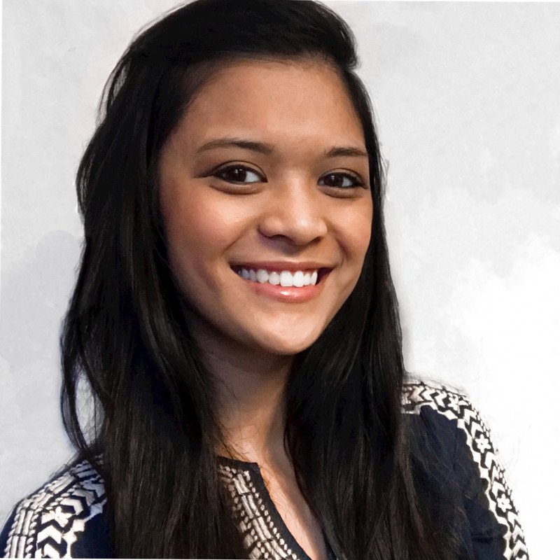 A young woman with long dark hair smiling with white teeth, sporting a patterned black and white top, standing against a plain white background.