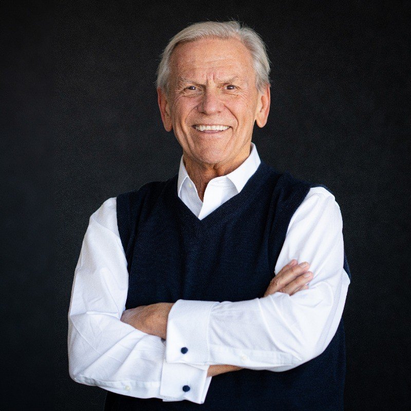An elderly man with gray hair smiling, wearing a white shirt and dark sleeveless sweater, crossing his arms against a dark background.