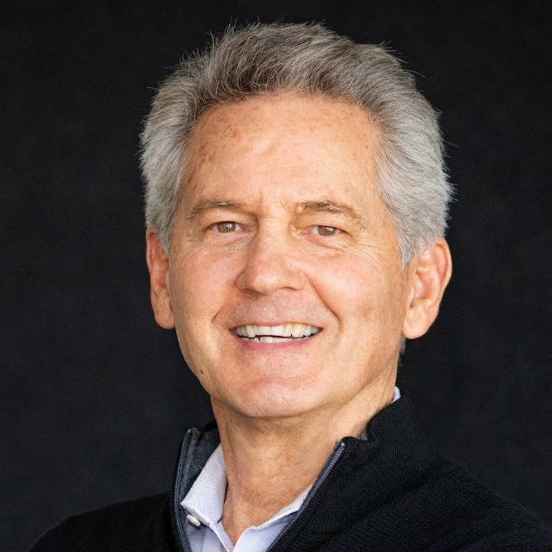 Portrait of an older man with gray hair, smiling, wearing a black zip-up jacket and a collared shirt against a black background.