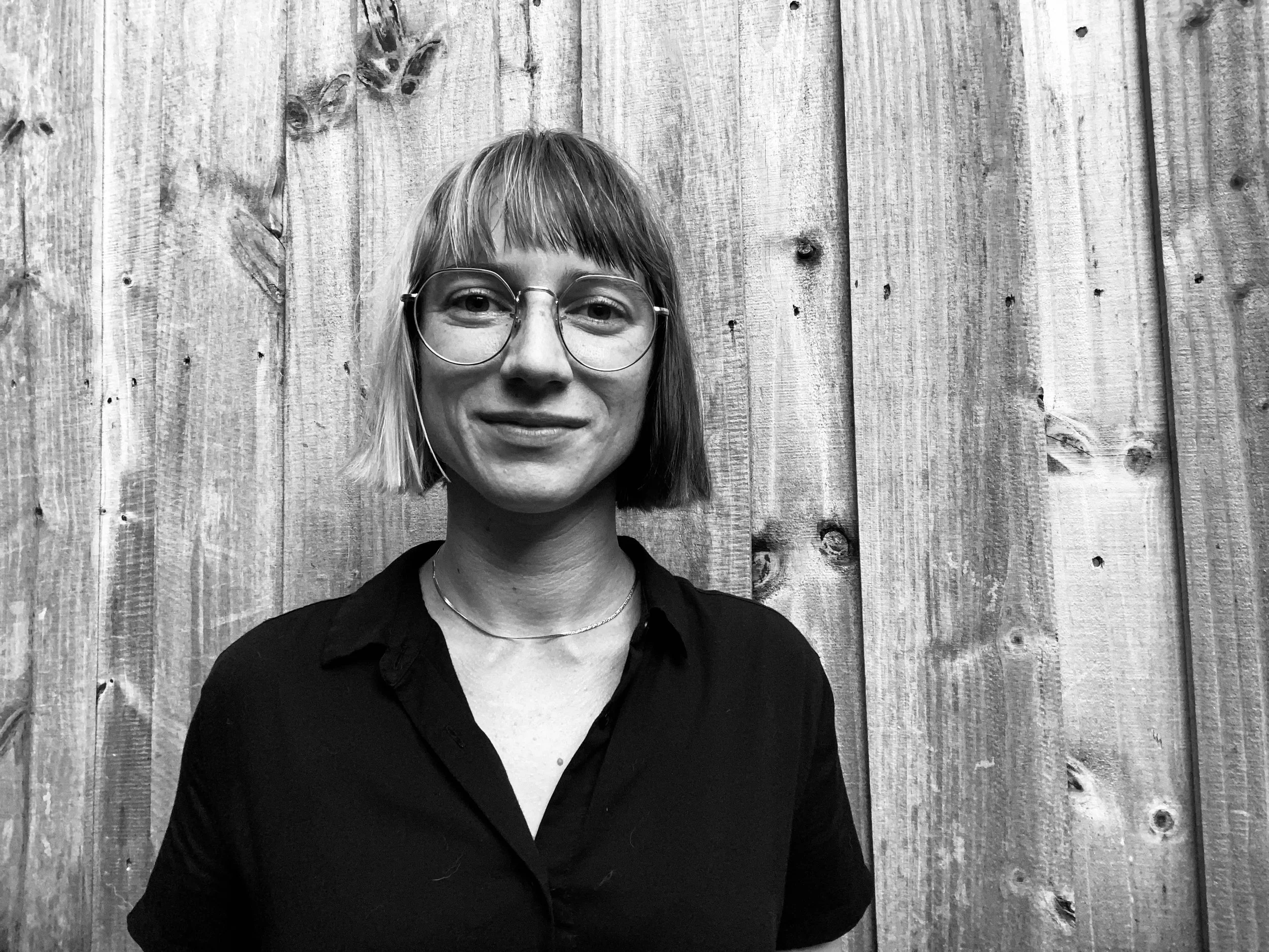 A woman with short hair, glasses, and a necklace standing in front of a wooden fence, smiling at the camera.