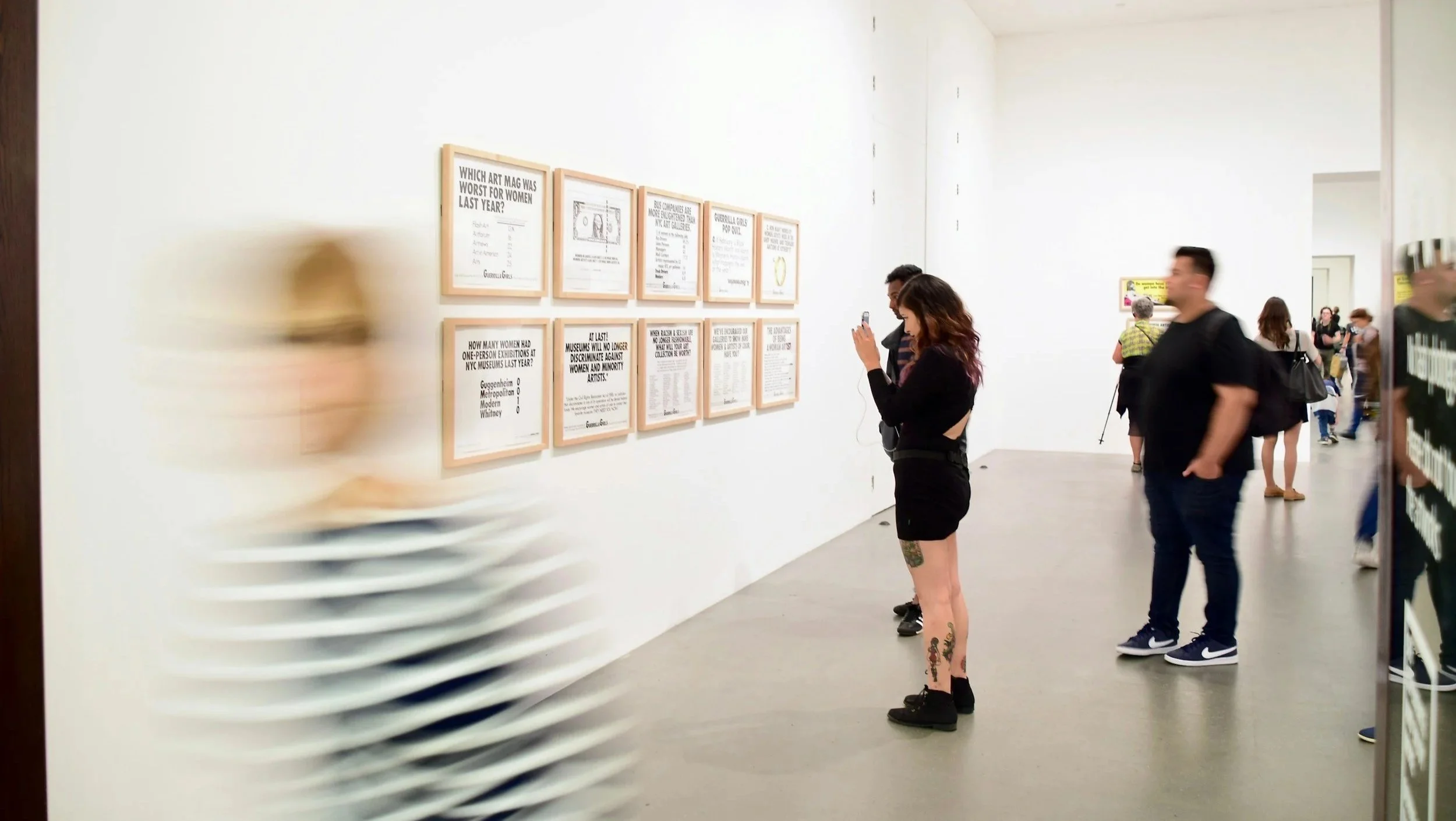 People viewing framed artworks and taking photos at an art gallery with white walls and a bright interior, some blurred figures in the foreground.