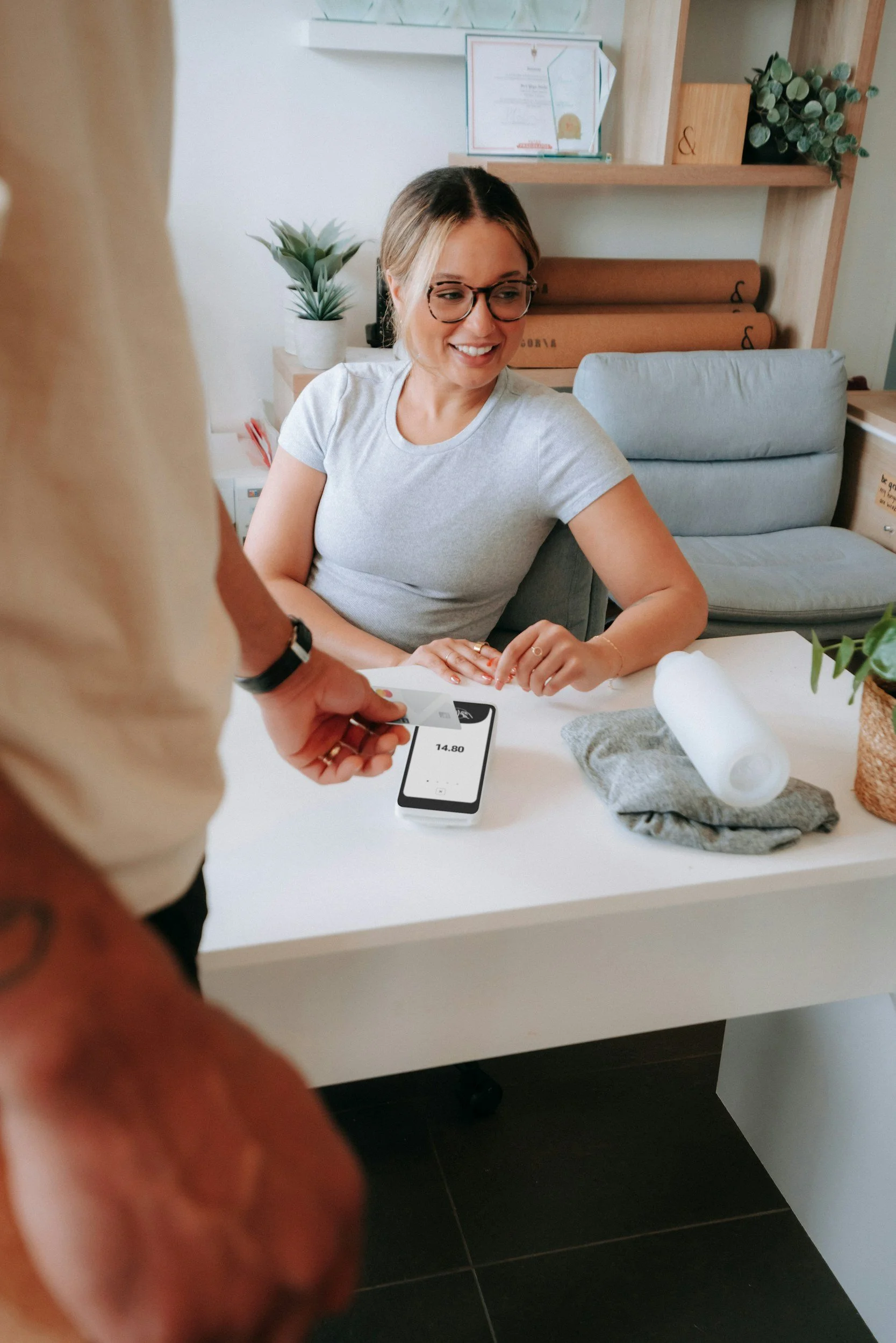 A woman is smiling while sitting at a white desk, paying with a credit card on a smartphone, with a person standing in front of her holding a card. The desk has a gray cloth, a roll of paper towels, and a potted plant. Behind her are shelves with certificates and a green plant, and a gray chair is in the background.