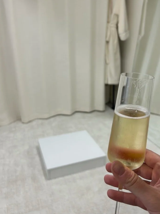 Hand holding a glass of champagne in a room with closed curtains and a white box on the floor.