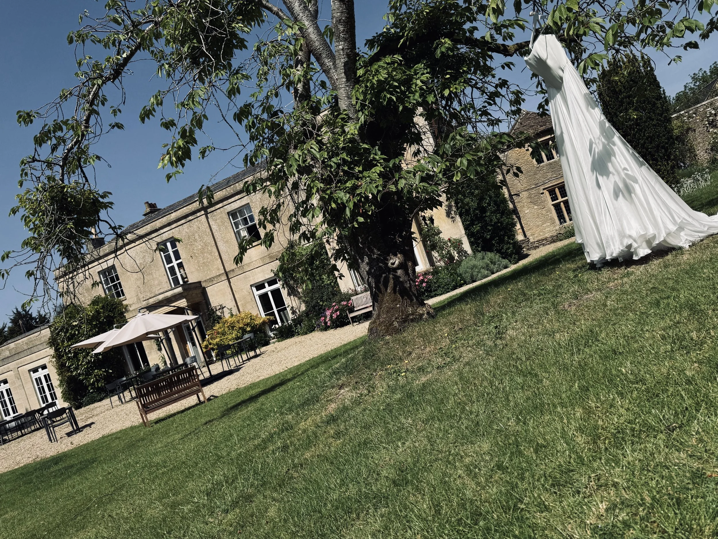 A white dress hanging on a tree branch in a garden with a stone building in the background under a clear blue sky.