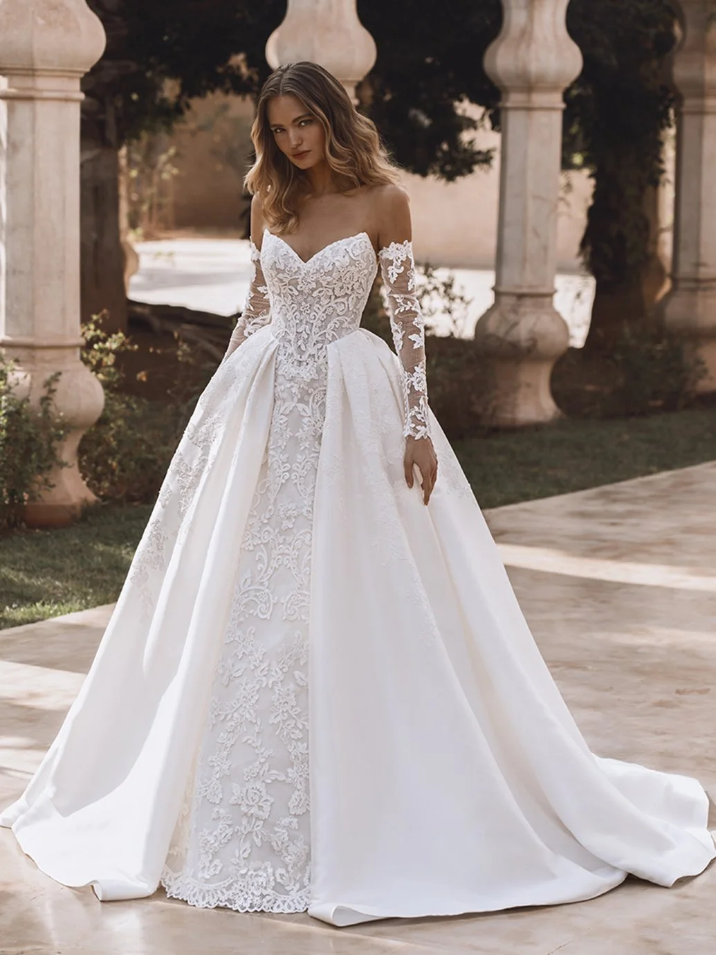 A bride in a lace couture gown with off-the-shoulder sleeves in a classical architectural setting.