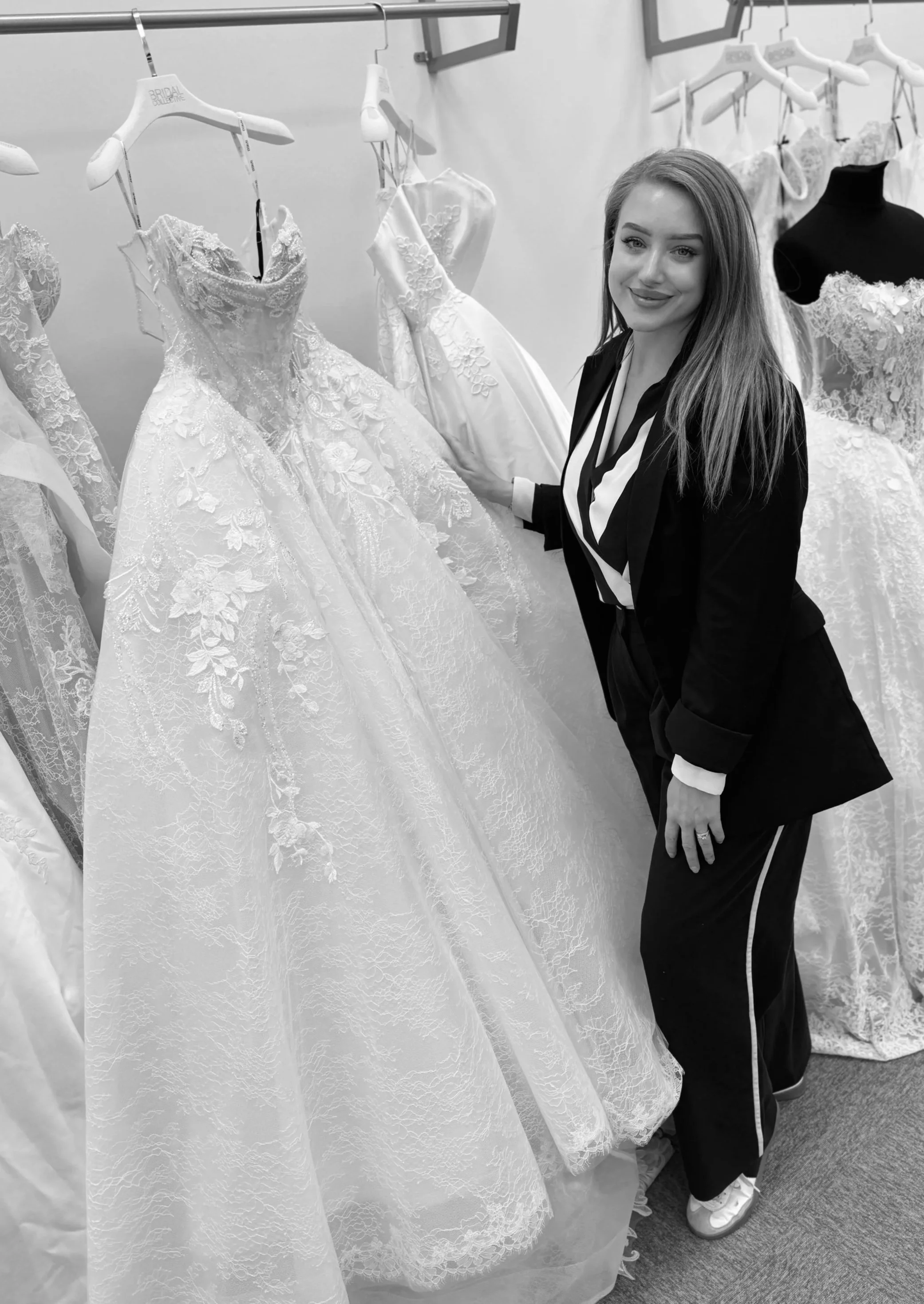 A woman wearing a blazer and track pants, smiling, shopping for wedding dresses in a store with multiple gowns hanging on racks.
