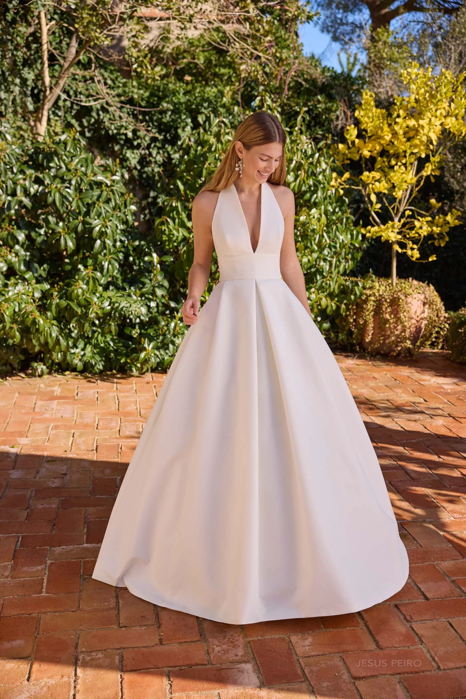 A woman in a white wedding dress standing outdoors on a brick patio with green and yellow foliage in the background, smiling and looking down.