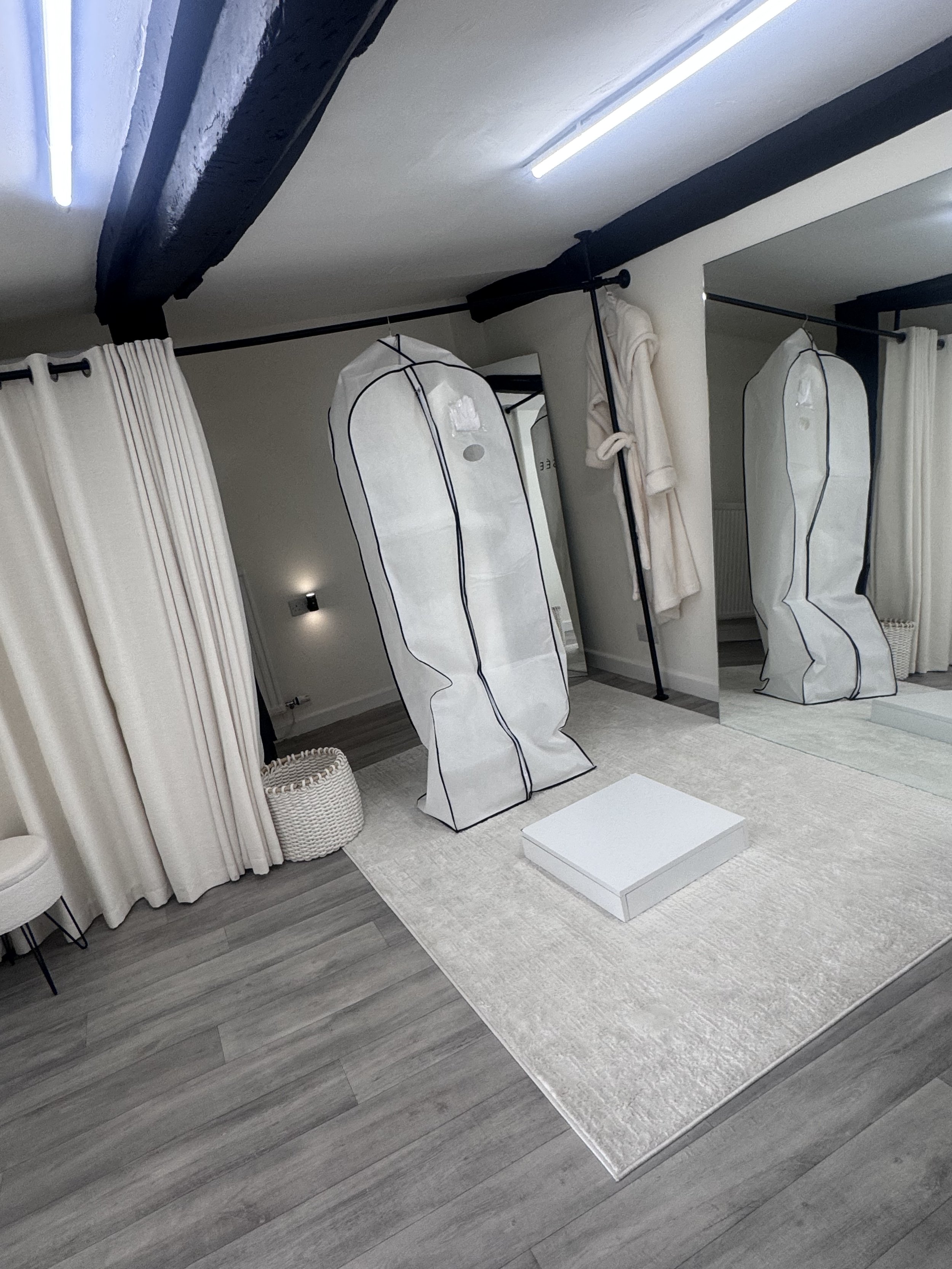 A fitting room area with white curtains, two white portable closet units, a beige bathrobe hanging, a mirror, a small white rug, and a white shoe box on a modern gray wood floor.