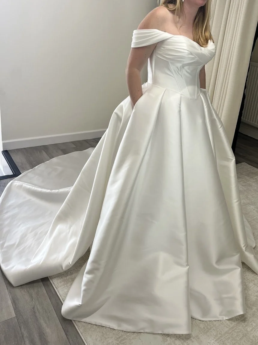 A woman wearing an elegant white off-shoulder wedding dress with a full skirt and train, standing indoors on a wooden floor near a mirror and light curtains.