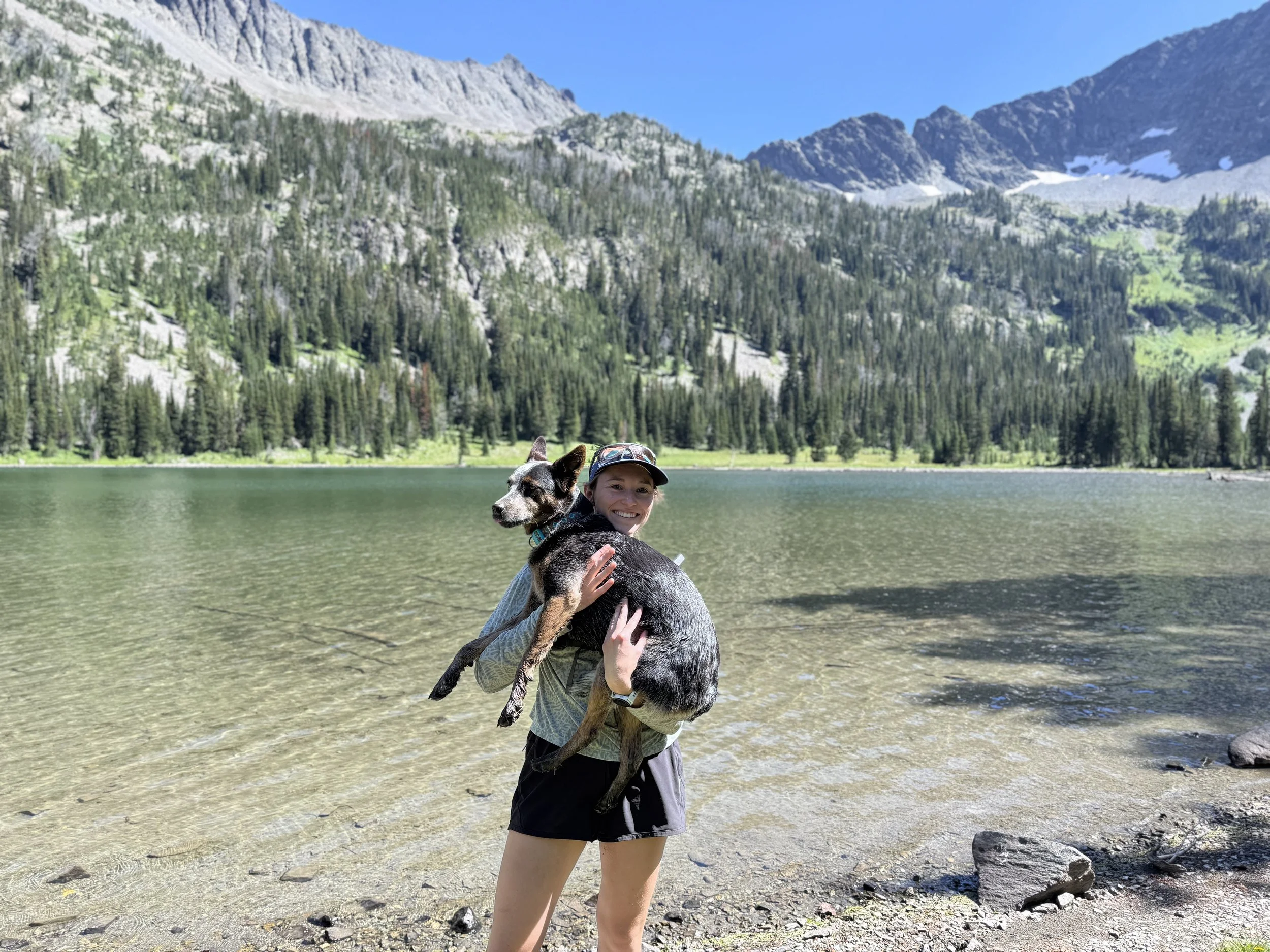 A woman smiling and holding a dog near a mountain lake with forested hills and rocky peaks in the background.