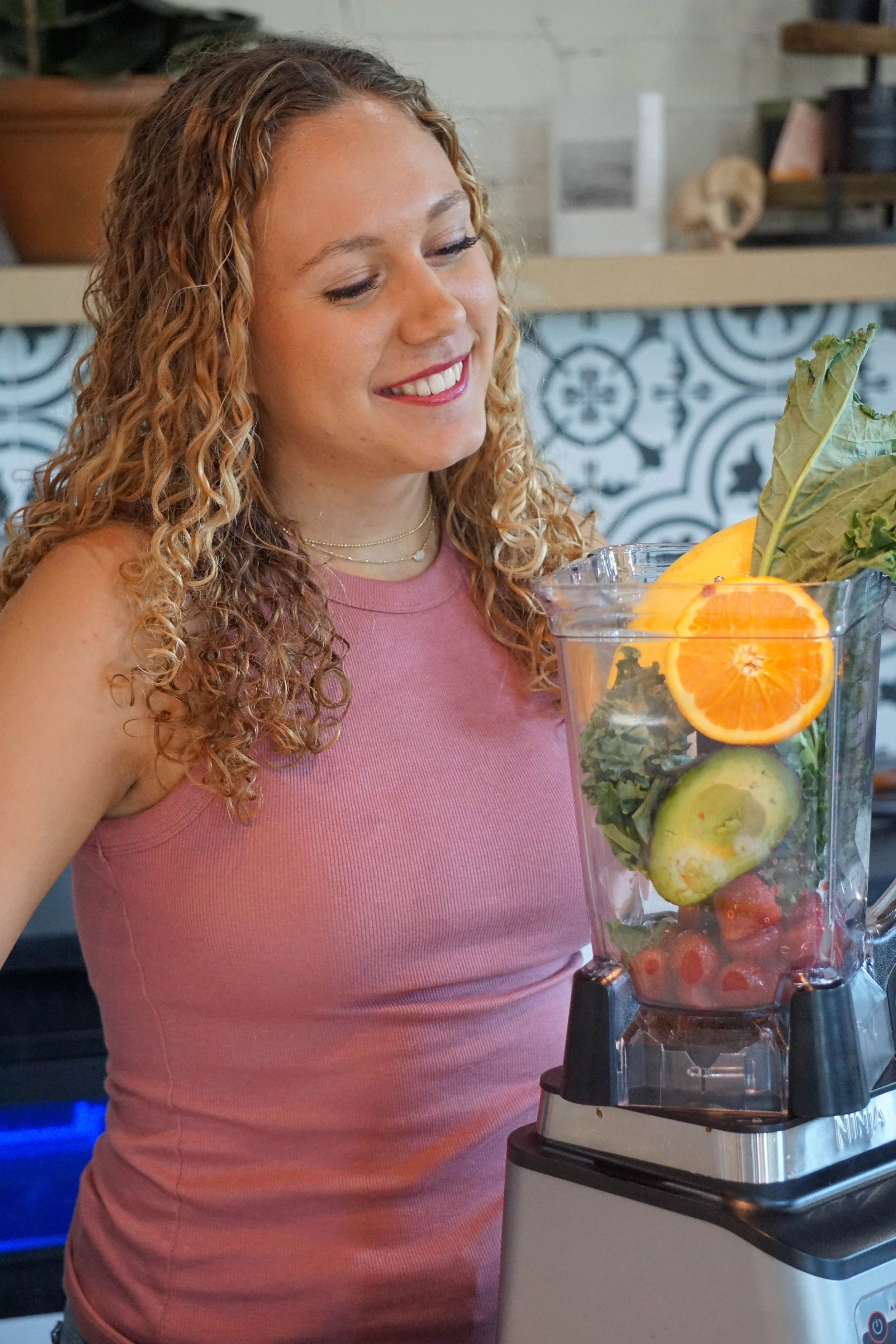 A woman smiling as she prepares a green smoothie with various fresh fruits and vegetables in a blender.