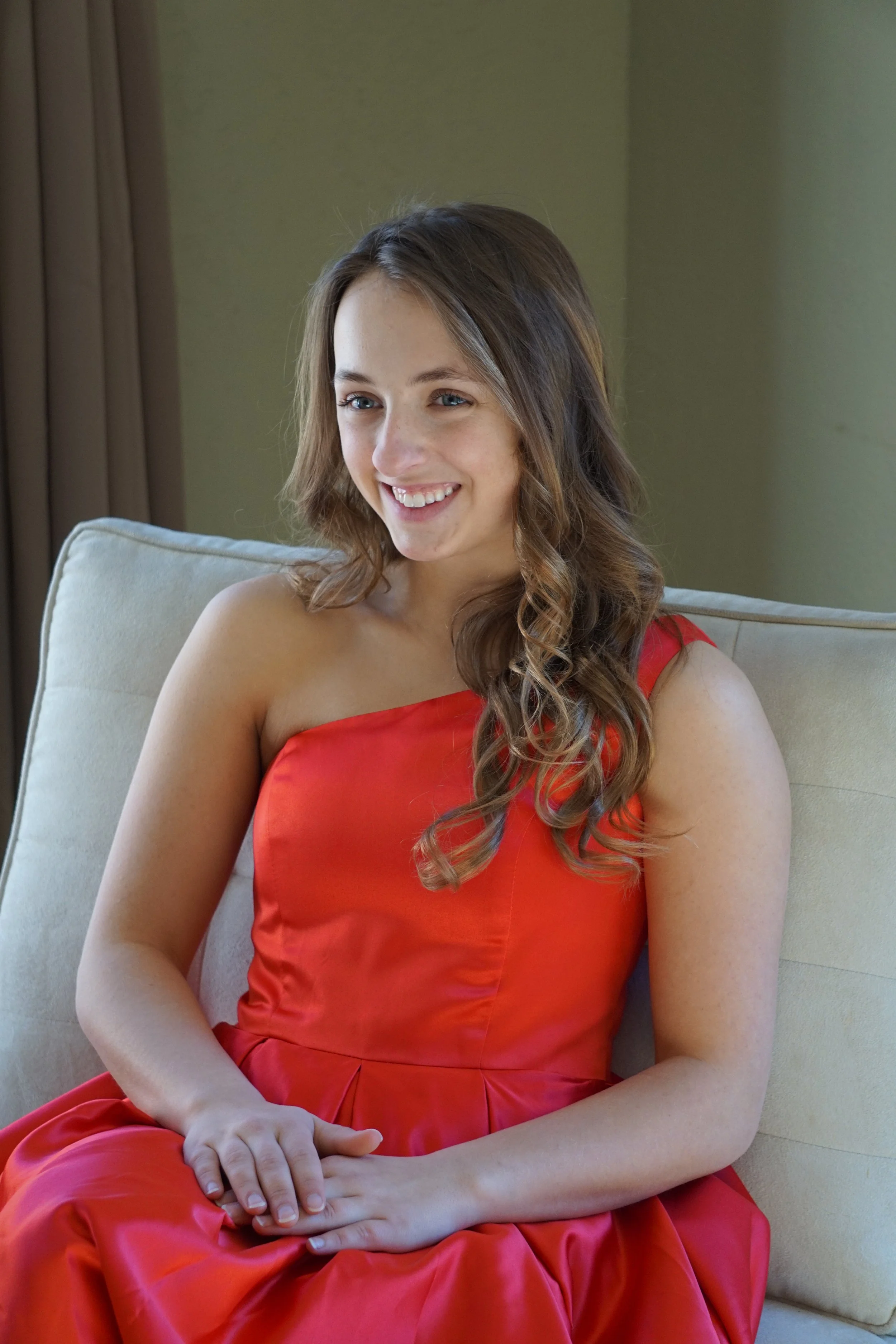 A young woman with long wavy brown hair, wearing a one-shoulder red dress, sitting on a beige sofa and smiling.