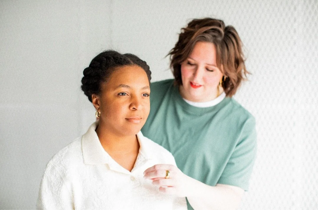 A woman with short, wavy hair in a green shirt gently touching a young woman with braided hair and earrings, who is looking forward.