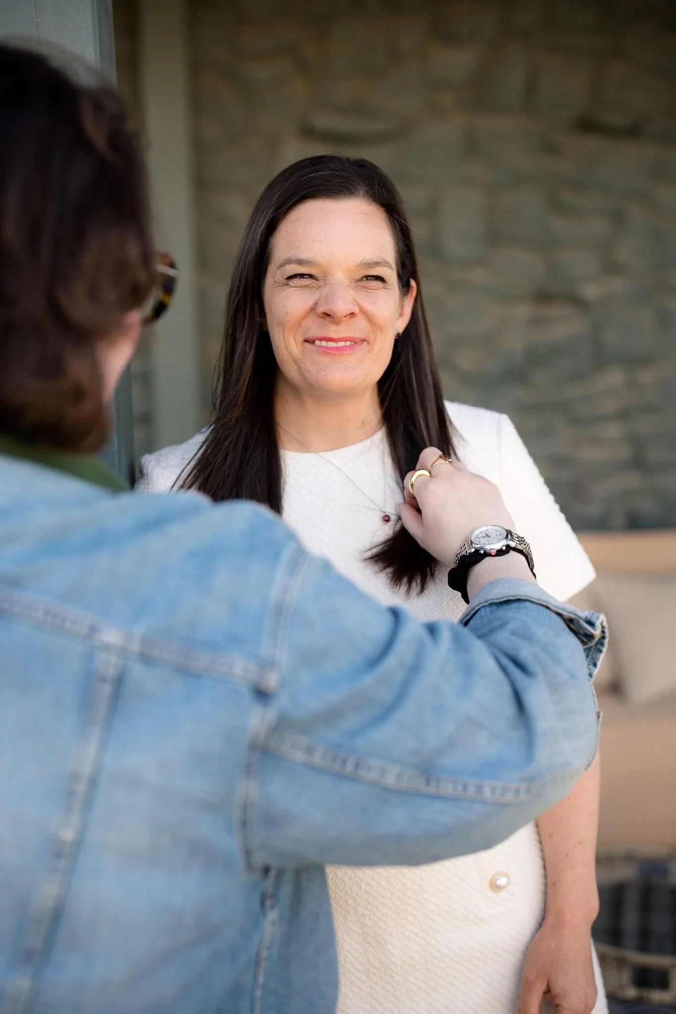 A woman with long dark hair and light skin smiling as another person adjusts a necklace on her. The woman is wearing a cream-colored top and is standing outdoors against a stone wall.