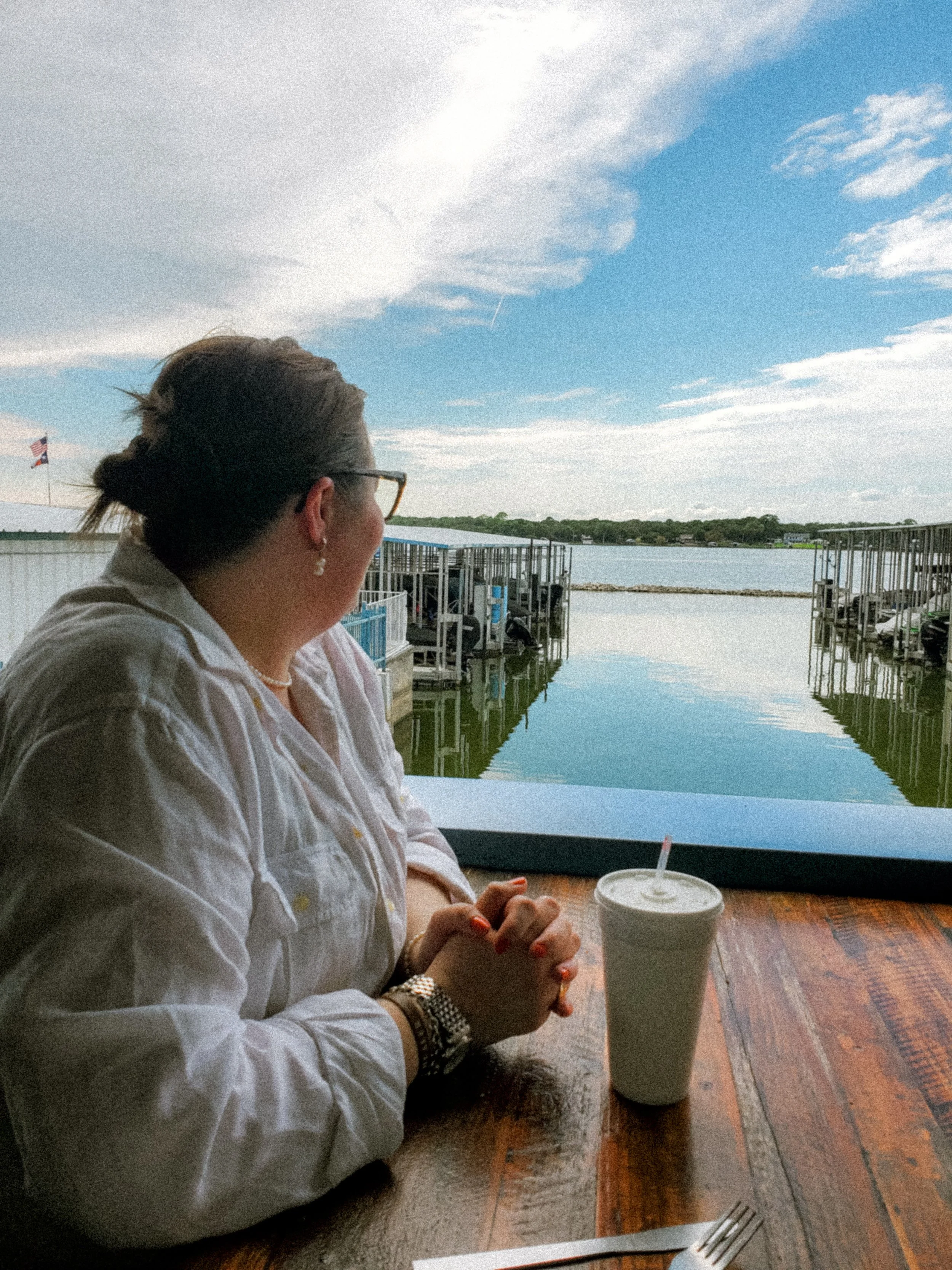 A woman sitting at a wooden table near a waterfront, gazing out over the water with a drink in a styrofoam cup in front of her. The water is calm, with docks and boats visible, and the sky is partly cloudy.