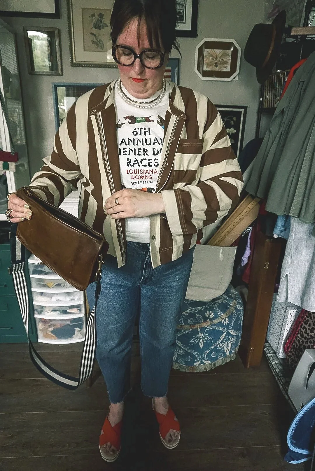 A woman standing indoors, wearing glasses, a striped brown and beige jacket, a white T-shirt with event details, blue jeans, orange wedge sandals, and holding a brown clutch purse with a striped strap.