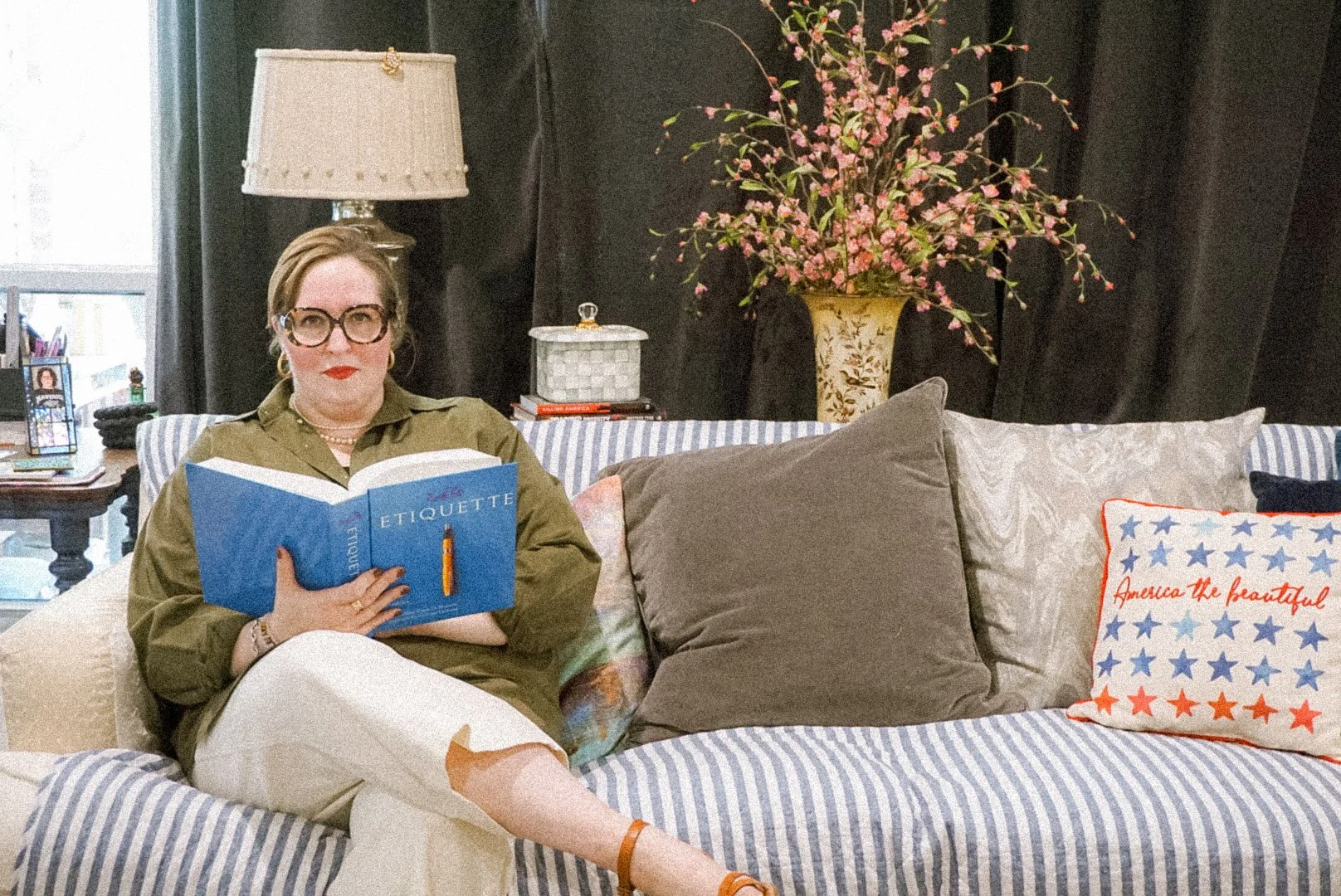 A woman with glasses and red lipstick sitting on a couch, reading a book titled "Etiquette." She is wearing an olive green shirt, cream pants, and tan sandals. The couch has striped cushions, and there are large pillows, one with stars and the phrase "America the beautiful," in front of her. Behind her is a side table with a lamp, a framed photo, and other items. There is a large vase with pink flowers, and black curtains in the background.