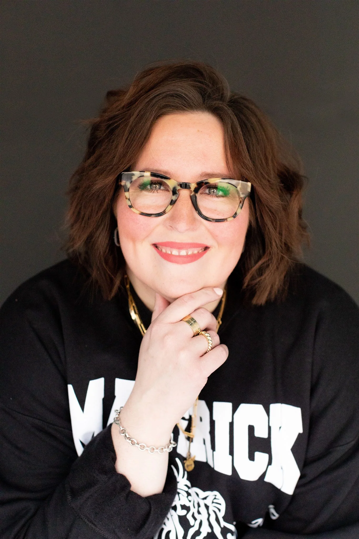 Portrait of a woman with glasses smiling and resting her chin on her hand, wearing a black sweatshirt with white text and jewelry, against a dark background.