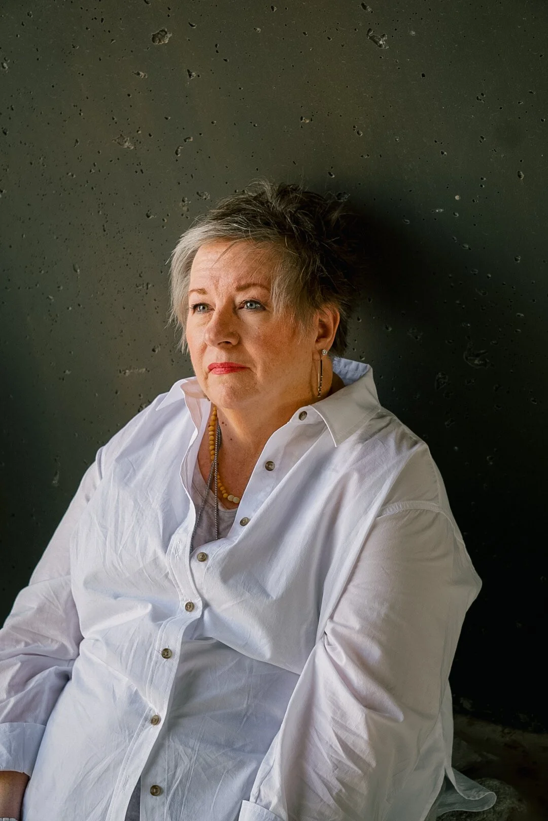 A woman with short gray hair sitting against a textured dark green wall, wearing a white button-up shirt and jewelry, looking to the side.