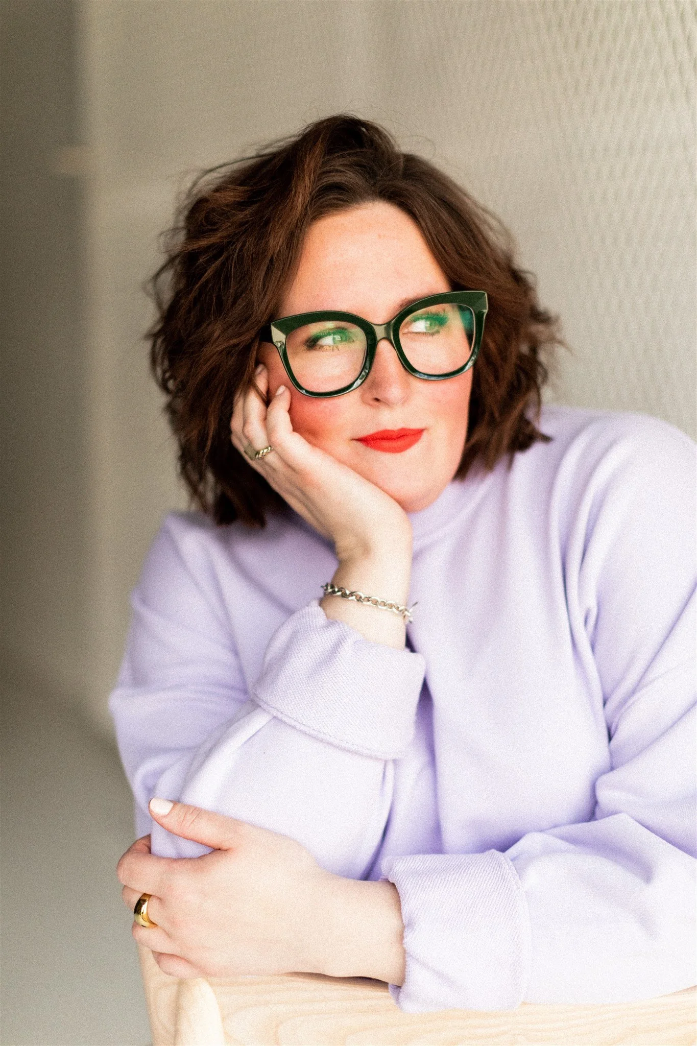 A woman with red lipstick, curly brown hair, black glasses, wearing a light purple sweater, and jewelry, resting her chin on her hand and looking out of the frame.