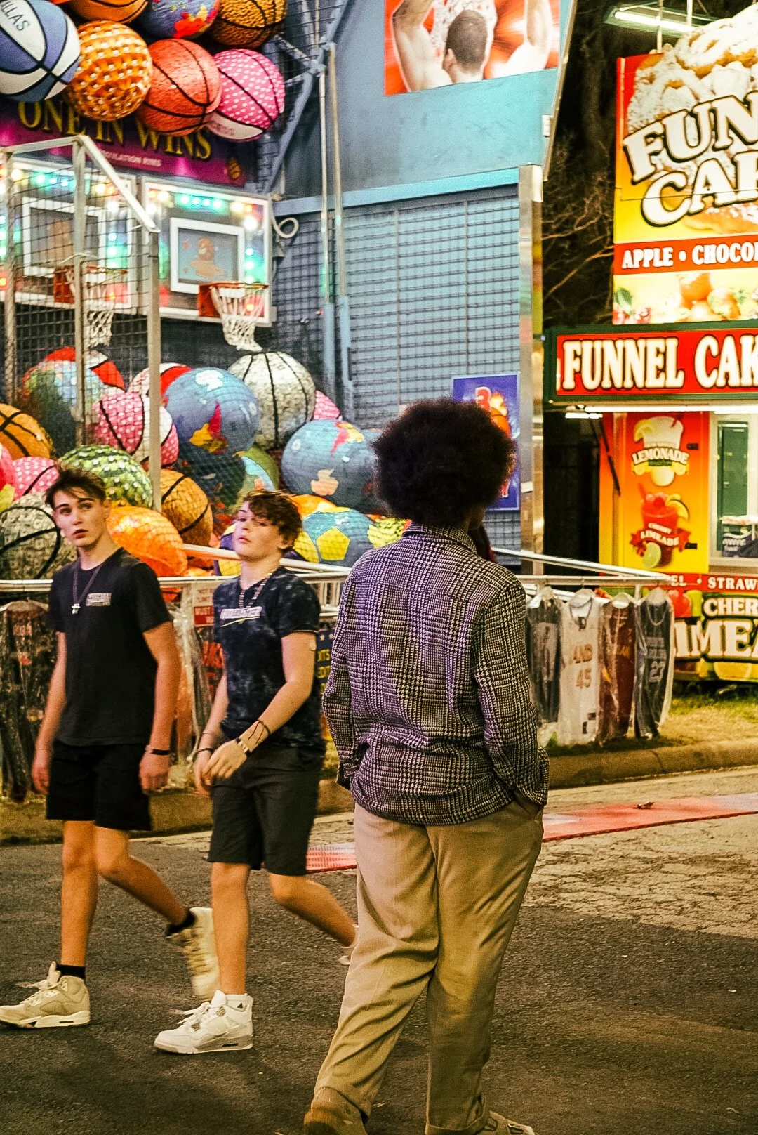 People walking past a carnival game booth with basketballs and colorful sports balls, signs advertising funnel cake and lemonade, and a bright, bustling fair atmosphere.