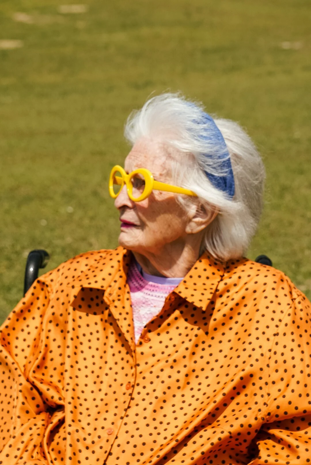 An elderly woman wearing yellow novelty glasses, an orange polka dot shirt, and a pink top underneath, sitting outdoors on a grassy area.