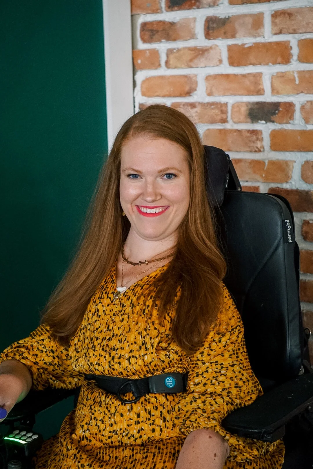 A woman with long red hair, wearing a yellow patterned dress and a necklace, smiling in front of a brick wall and green door.
