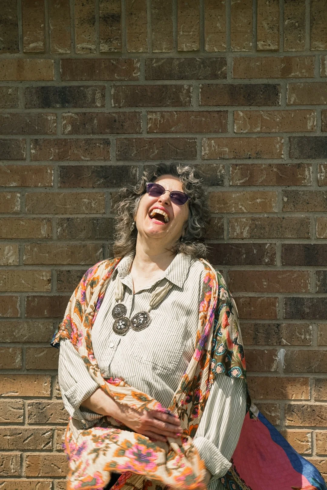 A woman with curly gray hair, wearing purple sunglasses, a cream striped shirt, a colorful floral scarf, and large decorative necklace, laughing outdoors against a brick wall.