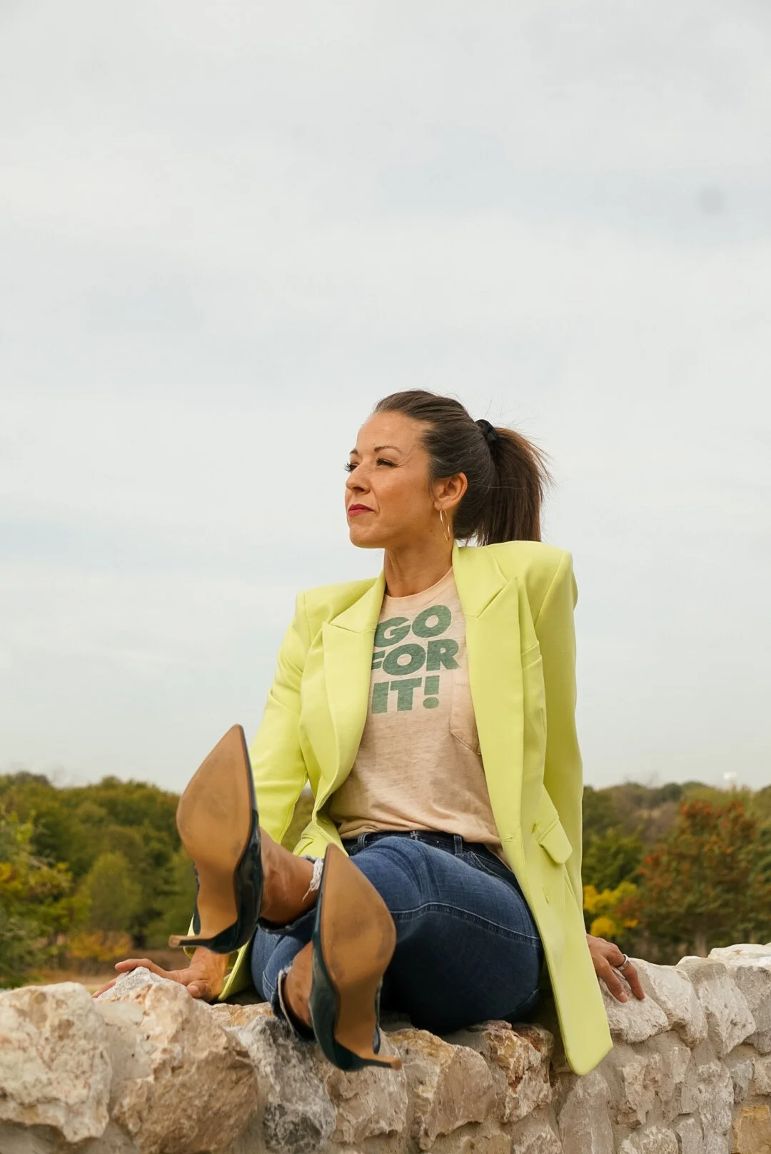 A woman sitting on a stone wall outdoors, wearing a yellow blazer, a beige T-shirt with green text, and jeans, with an expression of confidence.
