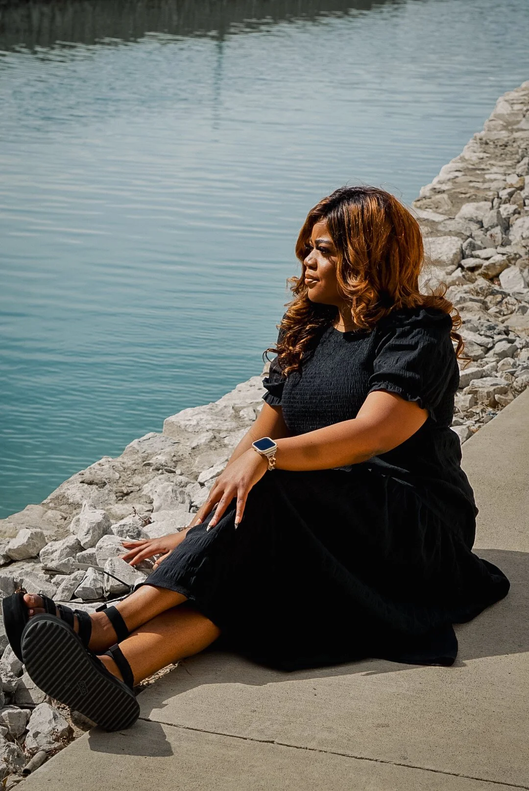 A woman with brown, curly hair, wearing a black dress, sitting on a concrete path next to a body of water, with rocks along the shoreline.