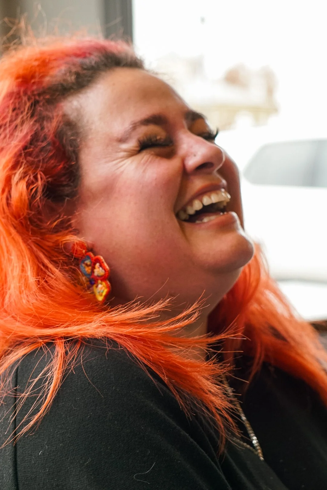 Close-up of a woman with bright orange hair, wearing colorful earrings, laughing with her eyes closed.