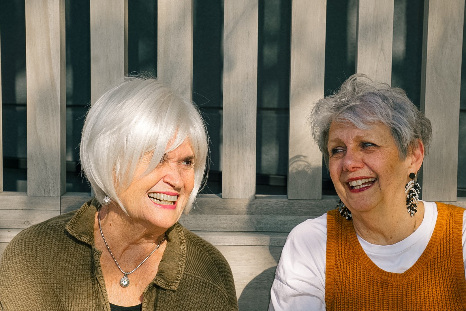 Two elderly women sitting outdoors and smiling at each other.