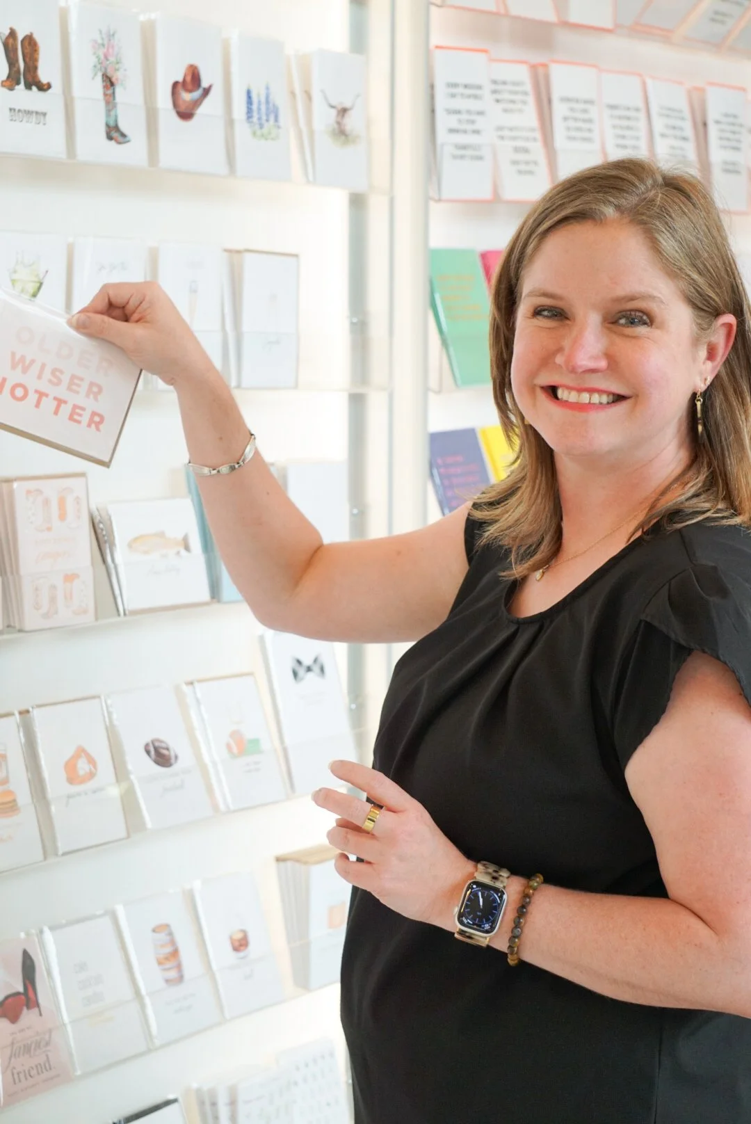A woman with shoulder-length brown hair, wearing a black sleeveless top, smiling, and pointing at greeting cards on a display wall in a store.