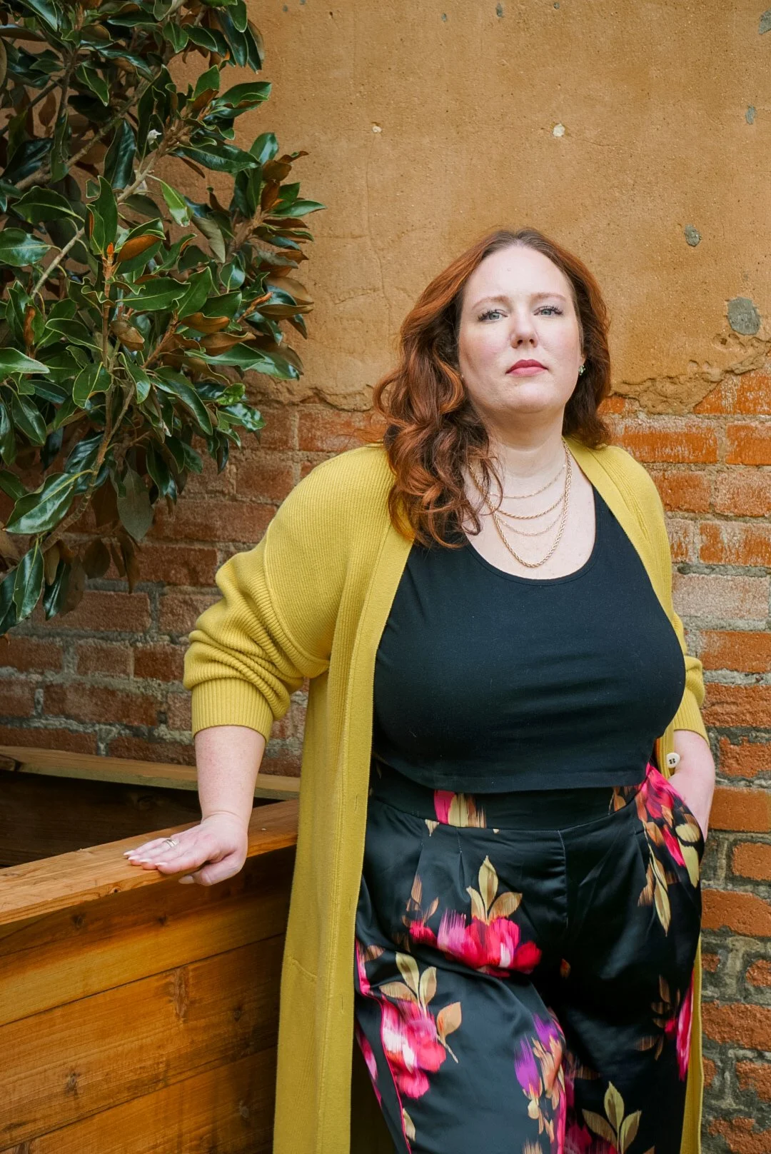 A woman with curly red hair standing against a brick wall and a potted plant. She wears a black top, floral pants, a yellow cardigan, and jewelry.