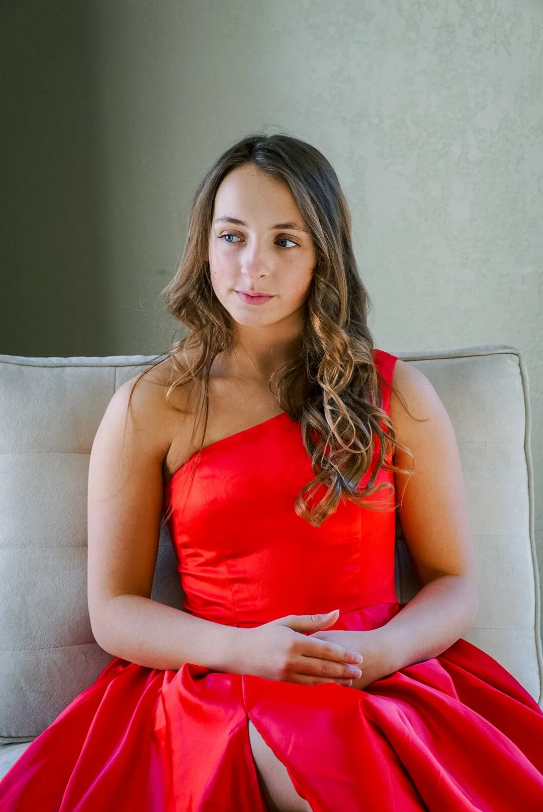 Young woman with wavy brown hair wearing a red dress, sitting on a beige sofa, looking thoughtfully to her left, in a softly lit room with neutral wall background.