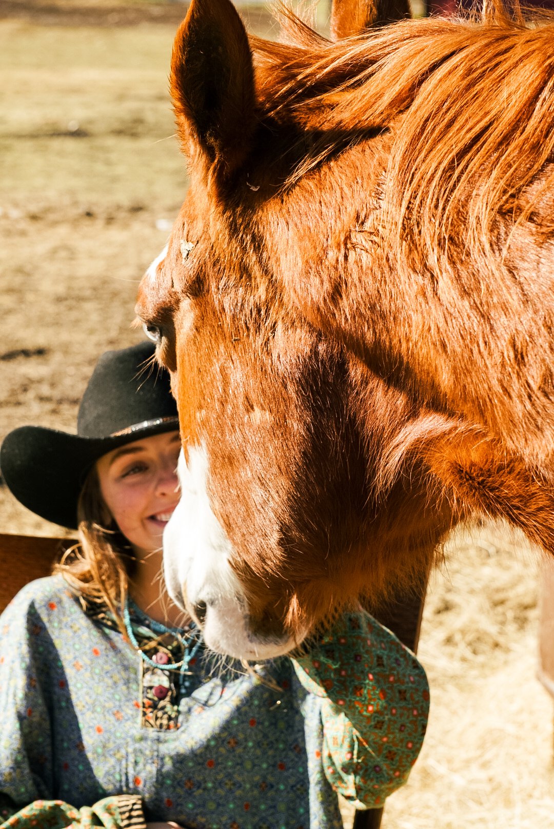 A young girl wearing a black cowboy hat and patterned shirt smiling as she interacts with a large brown horse with a white blaze on its face, in an outdoor setting.
