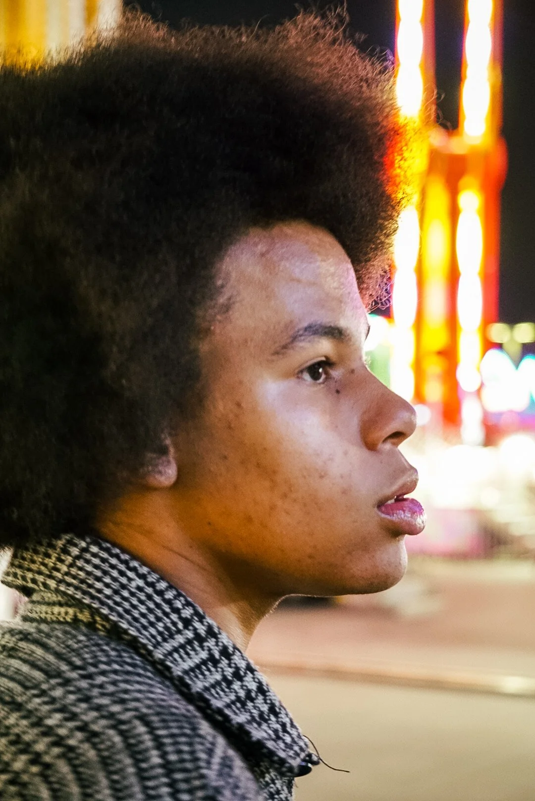 Close-up side profile of a young woman with natural hair and a patterned shirt, with neon lights blurred in the background.
