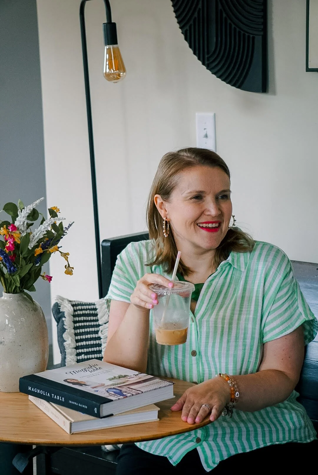 A woman with shoulder-length brown hair, wearing a green and white striped shirt, sitting at a wooden table with a book titled 'Magnolia Table' by Joanna Gaines, holding a plastic cup with a straw and a beverage inside, smiling in a cozy indoor setti