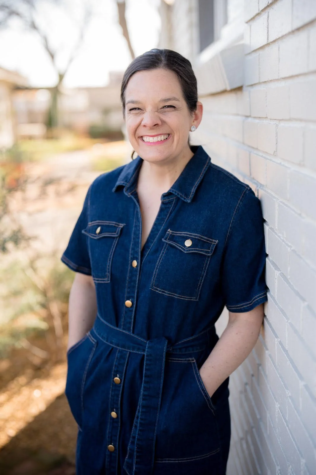 A woman with dark hair smiling and leaning against a white brick wall outdoors on a sunny day.