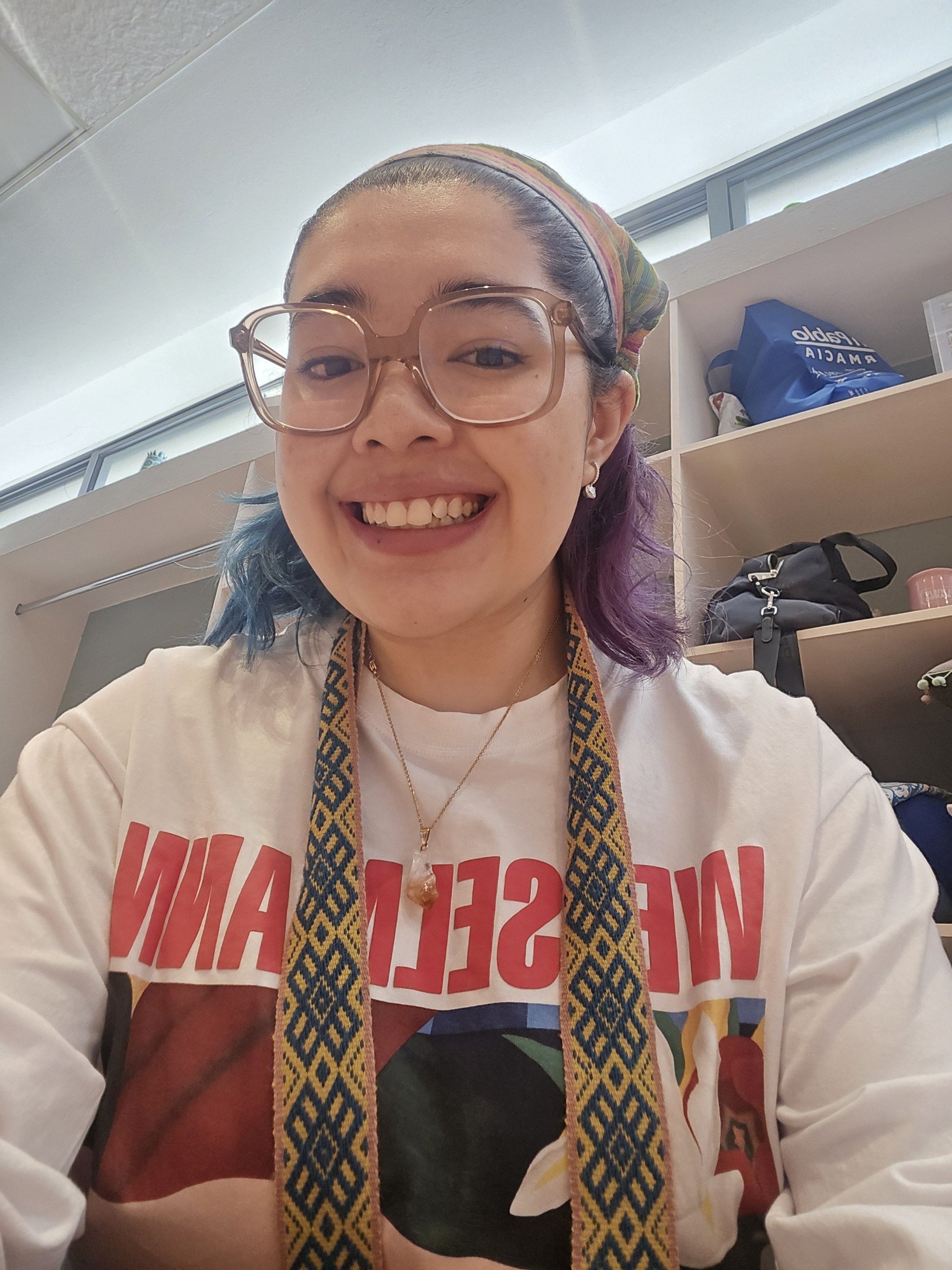 Una mujer sonriente con gafas grandes, usa una camiseta blanca con letras rojas y un collar brillante, lleva un pañuelo en la cabeza con colores y un cinturón de tela con patrón. Está en un lugar con estantes que contienen bolsas y otros objetos.