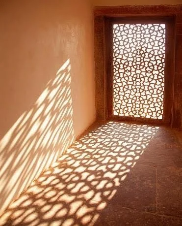 Sunlight passing through a patterned window grille, casting shadows across an interior wall and floor.