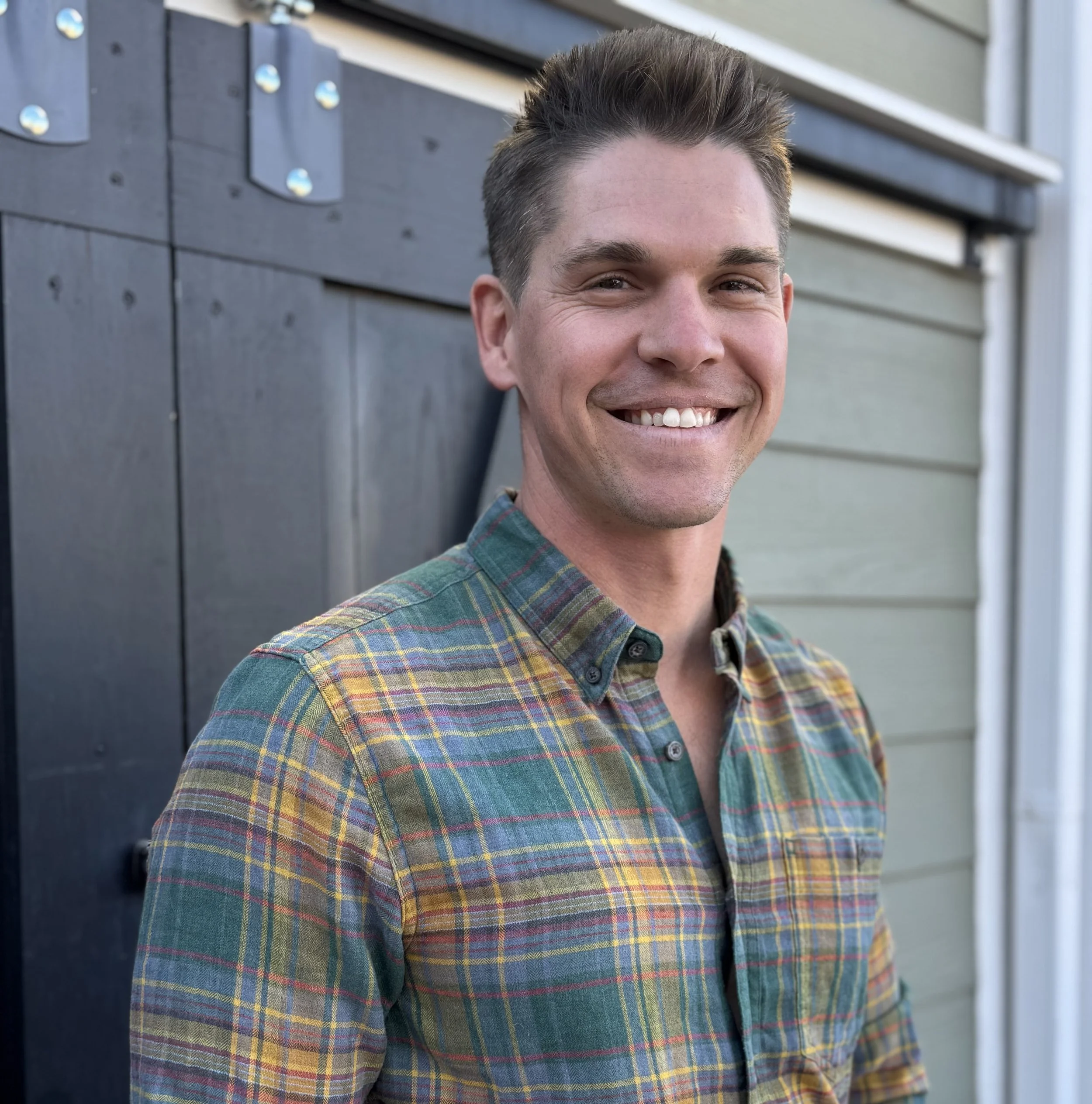 A young man with short dark hair and light skin, smiling in front of a yellow wooden wall, wearing a Patagonia fleece jacket.