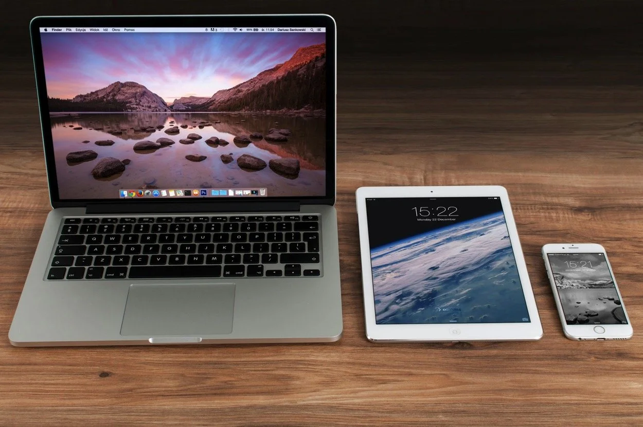 Open MacBook, iPad, and iPhone on a wooden table showing different screens and clocks.