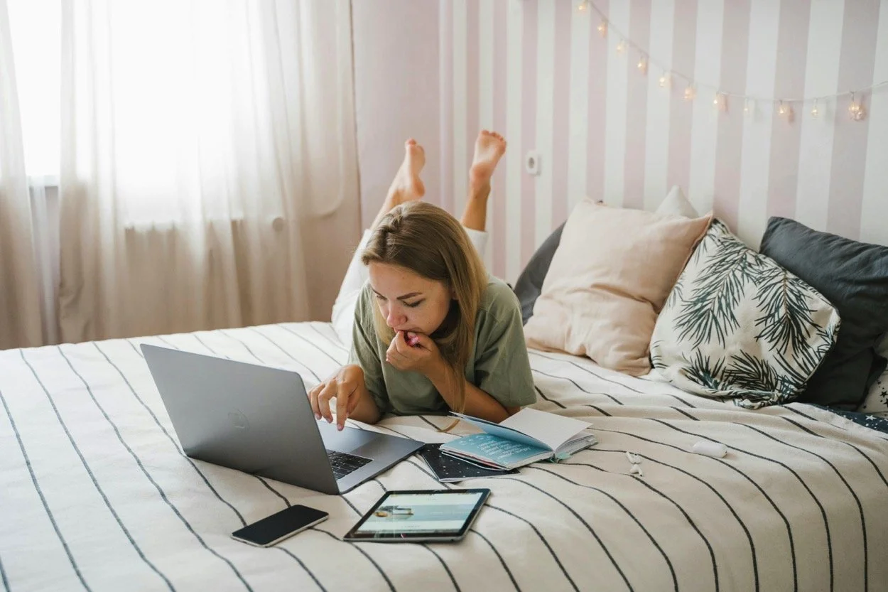 A young woman lying on a bed, working on a laptop, with a smartphone, tablet, and open notebooks nearby, in a bedroom with light-colored walls and string lights.