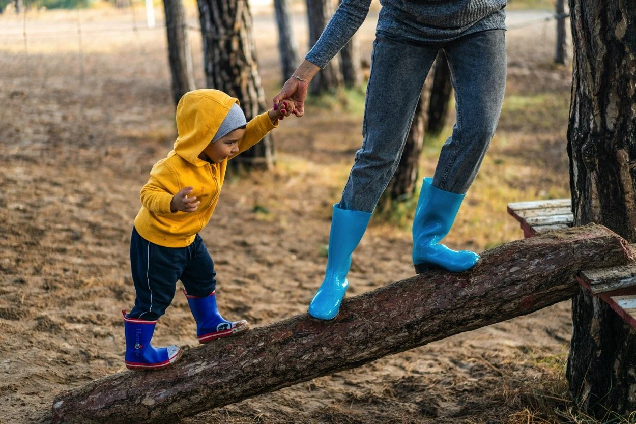 A young child wearing a yellow hoodie, blue pants, and rain boots, holding hands with an adult who is balancing on a log. The adult is wearing blue rain boots and grey jeans. They are outdoors in a wooded area with trees and dirt ground.