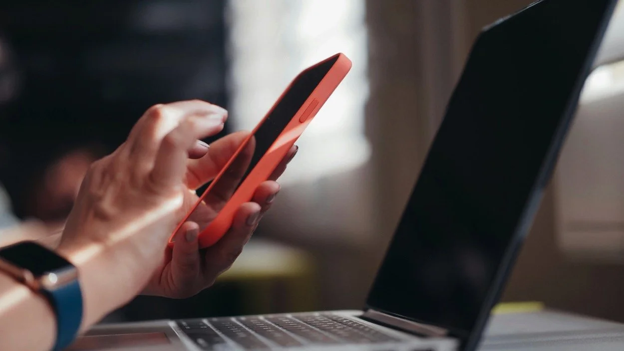 Close-up of a person's hand holding a smartphone over a laptop on a desk.