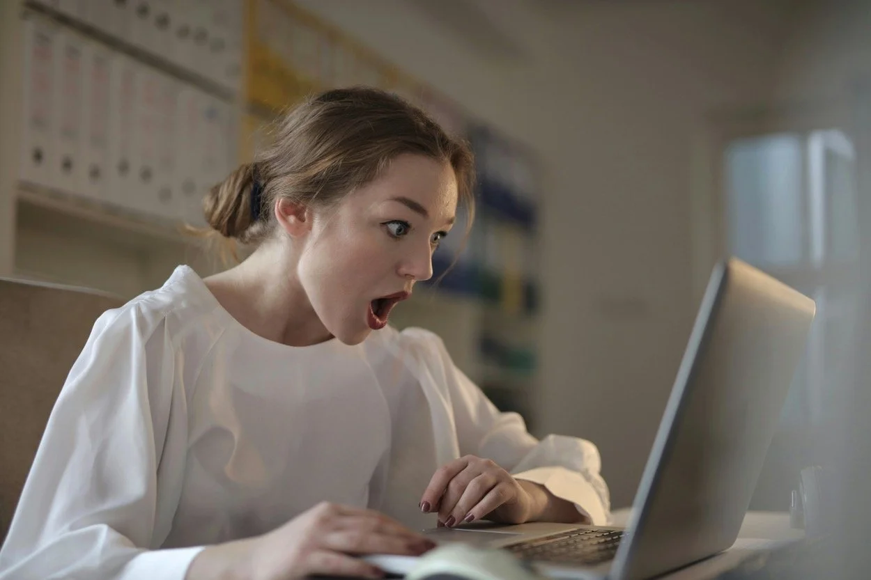 A woman in a white shirt with an open mouth and surprised expression, looking at a laptop screen.